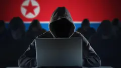 Uma pessoa com capuz e um computadorsafe online casino canadafrente a uma bandeira norte-coreana