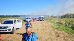 Щонайменше 11 людей у червоній формі з блакитними жилетками гуманітарної місії рухаються польовою дорогою далі від диму, який простягнувся на кількадесят метрів. Обабіч дороги стоять чотири білі автівки, в щонайменше двох вибиті вікна. На траві видно червоні плями – ймовірно елементи фірмового одягу