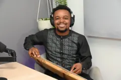 Déo Munyakazi yicaye mu ntebe y'umusego wa khaki arimo kumwenyura, afite inanga. Arareba muri 'camera', yambaye ishati y'umukara y'amaboko maremare irimo andi mabara yijimye, n'ipantalo y'umukara. Ku matwi yambaye ibikoresho bimufasha kumva (écouteurs).