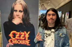 Um fã fazendo o sinal de rock com a mão, em pé ao lado de uma grande foto de Ozzy Osbourne