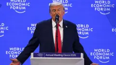 Kırmızı kravatlı lacivert takım elbiseli adam kürsüde konuşuyor. Arkasındaki mavi ekranda World Economic Forum yazıyor. 