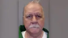 The prisoner is seen in a green jacket with a white shirt. He has a white moustache and white hair and there is a grey background