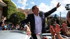 Jorge Tuto Quiroga, com jaqueta escura e camisa branca, acena do alto de um carro. Ao redor, veem-se mãos, câmeras fotográficas e, ao fundo, uma montanha.