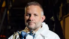 Fotografía de cerca de Ben Giles. Tiene 49 años y cabello corto y canoso. Lleva una mascarilla alrededor del cuello. Se puede ver su EPI, un traje blanco con capucha, alrededor del cuello.