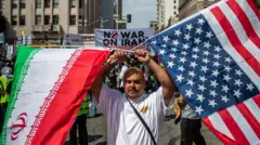 رجل طلب عدم الكشف عن هويته يحمل علماً إيرانياً وعلماً أمريكياً مقلوباً، بينما يتجمع الناس في فعالية "لا للحرب مع إيران وإسرائيل والمهاجرين" في 7 يونيو/حزيران 2025 في لوس أنجلوس، كاليفورنيا.
