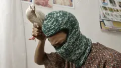 Foto de um homem inclinado para frente, olhando para baixo, enquanto segura uma pomba branca pelas pernas, acima da cabeça. Ele veste um balaclava de lã verde e uma camisa marrom estampada de manga curta