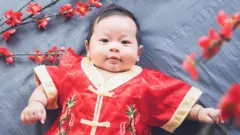 Bebê deitado sobre um tecido cinza, vestido com roupas tradicionais chinesas de cor vermelha e detalhes dourados, com flores vermelhas à sua volta