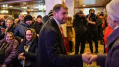 O candidato parlamentar da Moldávia, Ilan Shor, empresário, líder do partido que leva o seu nome e presidente da câmara da cidade de Orhei, encontra-se com apoiantes durante um evento de campanha na cidade de Comrat, em 15 de fevereiro de 2019. Ele veste um fato escuro e gravata vermelha e aperta a mão a alguém durante a campanha eleitoral. Várias senhoras idosas com lenços na cabeça estão ao fundo.