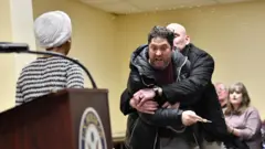 Ifoto igaragaza Ilhan Omar ari mu nama iruhande rw'indangururamajwi, ari imbere ahavugirwa ijambo. Imbere ye hari umugabo urimo kugundagurana n'undi amucigatiye mu maboko, ucigatiwe afite urushinje mu kiganza. Inyuma hirya haragaragara undi mugabo n'umugore bashungereye. 