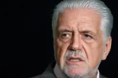 A imagem mostra Jaques Wagner em um retrato fechado, enquadrado dos ombros para cima, com cabelos brancos penteados para trás e barba curta grisalha. Ele olha para o lado, fora da câmera, com expressão séria e concentrada.