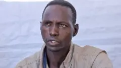 Retrato de um homem, do busto ao ombro, falando. Ele veste uma camisa listrada com gola. Um tecido azul pode ser visto atrás dele.