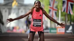 A etíope Tigst Assefa reage após cruzar a linha de chegada e vencer a prova feminina com um novo recorde mundial em campo exclusivamente feminino na Maratona de Londres de 2026. Ela está ajoelhada no chão, com os braços abertos.