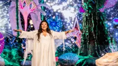 Regina Casé veste uma longa túnica branca e aparece no centro do palco com os braços abertos. Ao fundo, projeções coloridas de criaturas marinhas e formas orgânicas criam um cenário que remete aos primórdios da vida na Terra, complementado por elementos cenográficos que simulam rochas e vegetação.