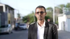 Homem negro com óculos escuros, terno preto e quipá na cabeça em bairro periférico