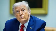 O presidente dos EUA, Donald Trump, sentado, com um terno azul, camisa azul, gravata vermelha e um broche na lapela com a bandeira do país