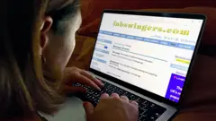 Mulher usa um laptop para acessar o site FabSwingers. Na tela, aparecem páginas de fóruns e mensagens. A imagem foi feita por trás do ombro da mulher, em um ambiente interno com pouca iluminação