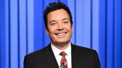 Jimmy Fallon, de terno em frente a uma cortina azul, pronuncia o monólogo de abertura do seu programa, na quinta-feira, 18 de setembro de 2025