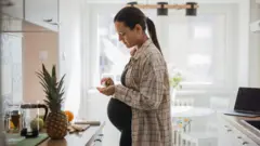 Mulher grávida, em pé, parada em frente ao balcão da cozinha, retirando uma cápsula de um frasco