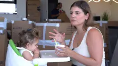 Uma mulher de cabelo castanho na altura dos ombros, vestindo uma blusa bege sem mangas, alimenta uma criança pequena sentada em uma cadeirinha. Ao fundo, há várias caixas de papelão.