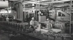 A imagem em preto e branco mostra uma linha de produção industrial dentro de uma fábrica ampla, com operários trabalhando em diferentes máquinas e equipamentos metálicos. Em primeiro plano, peças mecânicas alinhadas sobre uma esteira passam por processos de usinagem enquanto trabalhadores operam painéis de controle e acompanham a produção. O ambiente é iluminado por estruturas industriais no teto e tem aparência de registro histórico de uma fábrica automotiva ou metalúrgica.