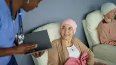 Paciente de câncer, com toalha na cabeça, sentada em uma poltrona, conversa com profissional de saúde