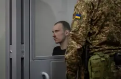 Dmitriy Kurashov, que tiene el pelo corto y viste una camiseta oscura, está sentado en el banquillo de los acusados, vigilado por un guardia ucraniano.