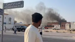 A man in a white shirt looks towards the scene of an Israeli airstrike on Doha. There is smoke rising in the background and people in the street in front of him who seem to be shouting and gesticulating