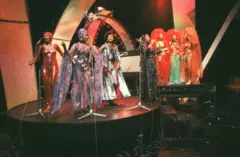 Uma apresentação musical vibrante com um grupo diversificado de artistas vestidos com trajes deslumbrantes com lantejoulas, capas e adornos ornamentados.