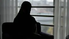 Amal aparece completamente recortada contra la luz, sentada en el interior junto a un gran ventanal con cortinas transparentes, con la mirada puesta en un paisaje urbano difuminado.