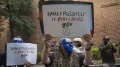 Manifestantes com máscaras azuis mostram cartazes com os dizeres: "Sorria, a Palantir está observando você"