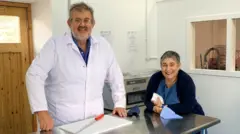 Richard Irvine de pie detrás de un mostrador metálico. Tiene el pelo y la barba canosos, y lleva gafas, una camisa azul rey y un abrigo blanco. Su esposa está a su lado, con un cárdigan azul marino, un vestido azul y un lápiz labial rojo oscuro. Tiene el pelo canoso y muy corto.
