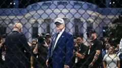 USAの文字が書かれた白い帽子をかぶったダークスーツ姿のトランプ氏が、金網越しに前方を見ている。背後にはテレビカメラをもった人などが立っていて、その後ろにホワイトハウスの建物が見える