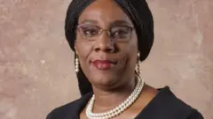 Dr. Olugbemisola Odusote wear black cloth wit cream colour beaded earrings and necklace. She wear eye glass and pack her black long braid for back.
She smile to di camera wit her maroon red lipstick.