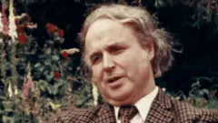 Noel Stanton veste um terno de tweed com corte de cabelo para disfarçar a calvície, em uma entrevista para a TV nos anos 1970 ou 80