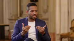 Rick Azevedo gesticulando durante entrevista à BBC. Ele veste terno azul e camiseta branca, e usa um cavanhaque e cabelo curto, raspado dos lados