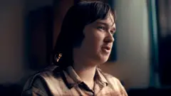 Calum, um jovem com cabelo castanho comprido e leve barba, vestindo uma camisa xadrez marrom, aparece olhando para a direita da câmera em um ambiente escuro, com as cortinas fechadas ao fundo e uma luz forte incidindo sobre seu rosto pelo lado direito da imagem