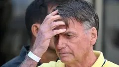 Jair Bolsonaro mirando hacia la izquierda y con la mano derecha sobre la frente. Viste una camiseta con cuello amarilla.