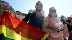 A couple show a rainbow flag and a toy that looks like Pope Leo