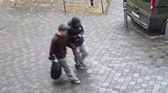 Imagen fija de una cámara de seguridad que muestra desde arriba a dos chicos adolescentes, vestidos de manera informal, caminando por una calle pavimentada. Sus rostros están borrosos. El que está más cerca de la cámara lleva una mochila negra en la mano derecha.