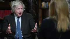 Former prime minister Boris Johnson speaking while holding out both hands. In the foreground, out of focus, we see the back of a woman with blonde hair.