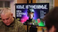 На переднем плане мужчина в камуфляже, на заднем, на большом экране, - реклама службы в беспилотных войсках