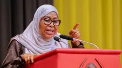 Perezida Samia Suluhu avugira imbere y'ikoraniro ry'abantu, yazamuye urutoki, yambaye umwenda w'ikigina n'igishura cy'umweru