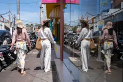 Dwie młode kobiety w letnich strojach i kapeluszach idą ulicą. Jedna z nich patrzy w witrynę sklepową. Na krawężniku rzędami zaparkowano skutery i samochody. Kuta, Bali, Indonezja. 5 marca 2026 r. 