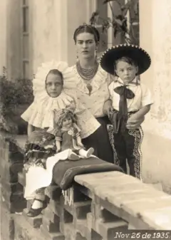 Frida, em pé ao centro, usa uma blusa branca com pequenas flores bordadas, uma saia escura, um colar de várias voltas e brincos longos. Ao seu lado está Antonio, em pé, usando camisa branca, chapéu e uma calça de mariachi com um laço preto no pescoço. Do outro lado de Frida, sentada, está Isolda, vestida com traje de tehuana e sapatos pretos. Uma boneca está sentada à frente delas. Estão em um jardim