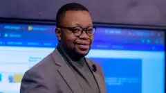 Jean Maurice Uwera yambaye indorerwamo, ikoti ririmo uturongo tw'ikigina, imbere harimo umupira w'icyatsi kibisi. Ku ikoti hometseho indangururamajwi nto yo kuvugiramo. Inyuma ye hari 'screen' nini iriho amabara y'ubururu, umweru n'umuhondo. Ibiyiriho ntibisomeka. 