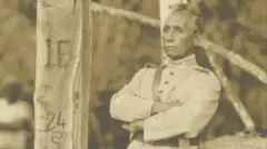 Foto em sépia mostra Rondon de braços cruzados olhando para o horizonte ao lado de uma árvore