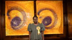 تصویر ننا کالو، هنرمند، که مقابل دو اثر طراحی خود با رنگ‌های زرد و بنفش ایستاده است