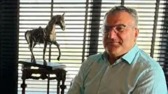 Cem Küçük pencere önünde iç mekanda oturuyor kameraya bakıyor, gönlekli. Geride bir at biblosu var. 