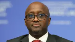 Minisitiri w'ububanyi n'amahanga w'u Rwanda Olivier Nduhungirehe yambaye ikositimu y'umukara, ishati y'umweru na karuvati y'umutuku irimo utubara tw'umukara. Arareba muri 'camera' asa nk'ureba hejuru gato. Yari yitabiriye inama ku mahoro n'uburumbuke mu karere k'ibiyaga bigari, yabereye i Paris mu Bufaransa, ku itariki ya 30 Ukwakira (10) mu 2025.