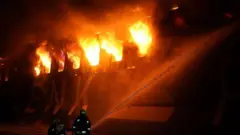 یک کشتی باری که آتش گرفته و چند آتش‌نشان مشغول اطفای حریق 